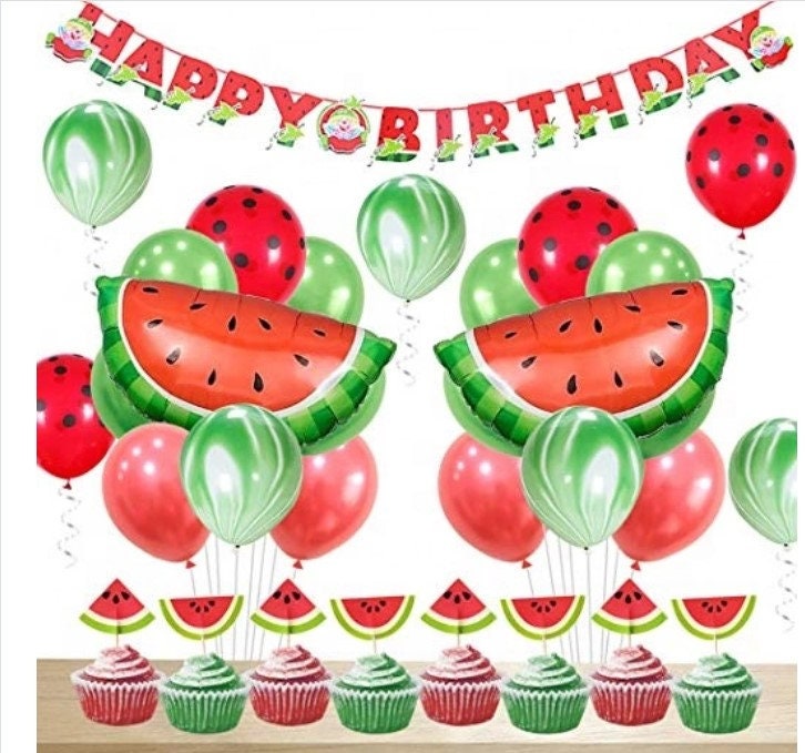 Assorted watermelon party supplies, including a banner, balloons, and cupcake toppers, laid out for a themed celebration.