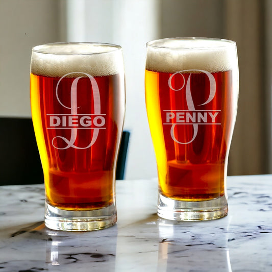 A clear monogrammed beer glass with an etched initial 'S' and a small design, standing on a wooden surface.
