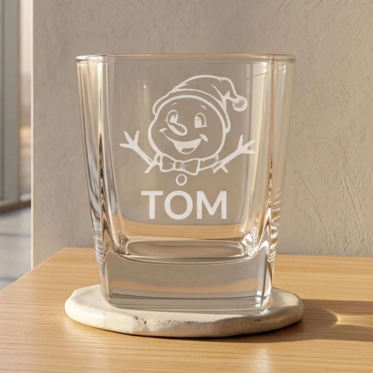 A clear square drinking glass with a frosted white front featuring a gray snowman face and the name 'TOM' in gray text, centered on a reflective surface with blurred Christmas trees and red ornaments in the background.