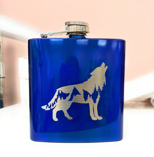 Close-up of a blue powder-coated hip flask with a wolf and pine forest engraving.
