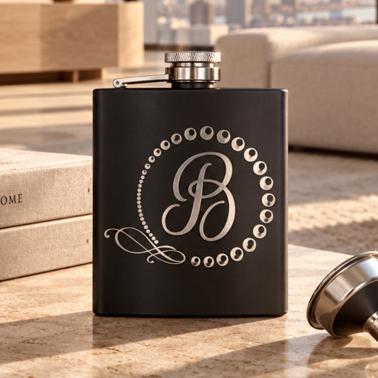 Black 6 oz stainless steel hip flask laser engraved with script monogram letter B in a dotted wreath, with funnel on a table