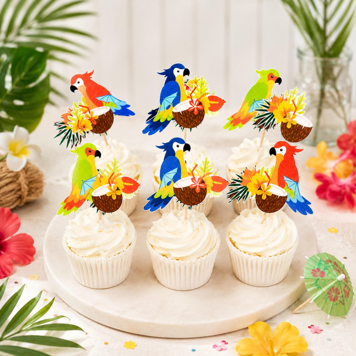 Six colorful parrot cupcake toppers arranged in white frosted cupcakes with palm leaves and tropical flowers.