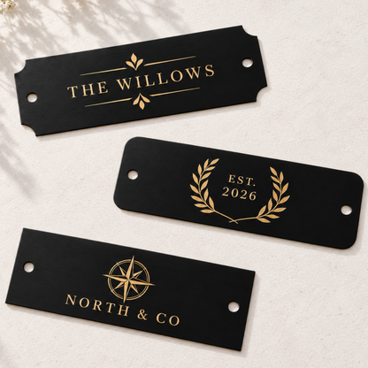 Three black rectangular personalized name plates with gold foil lettering and decorative graphics, arranged on a light textured surface with soft shadows and small white flowers.